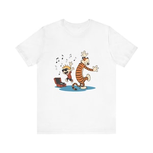 Puede incluir: Camiseta blanca con una ilustración de dibujos animados de un niño y un tigre bailando cerca de un tocadiscos. El niño lleva gafas de sol y el tigre tiene rayas naranjas y negras. Notas musicales flotan en el aire.