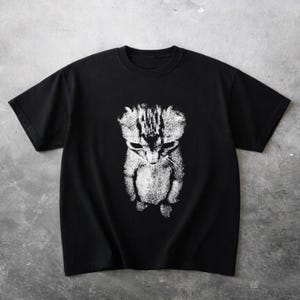 Könnte beinhalten: Schwarzes T-Shirt mit einem verblassten weißen Motiv einer traurig aussehenden Katze. Das Gesicht und der Körper der Katze sind in einem detaillierten, künstlerischen Stil dargestellt. Das T-Shirt hat kurze Ärmel und einen Rundhalsausschnitt, geeignet für Freizeitkleidung.