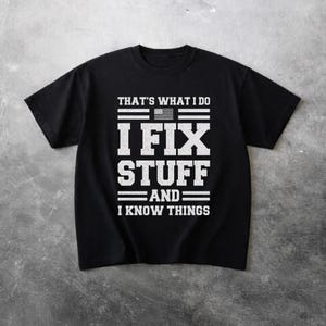Puede incluir: Camiseta negra con el texto blanco "THAT'S WHAT I DO, I FIX STUFF AND I KNOW THINGS". El diseño incluye una bandera estadounidense encima de las palabras "I FIX STUFF".