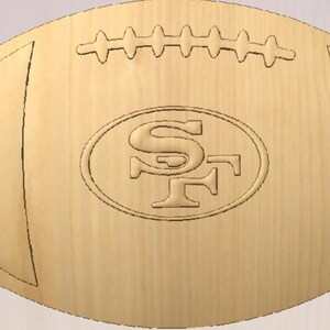 May include: A wooden football with the San Francisco 49ers logo carved into the center. The football is light brown with carved details, including the laces and the team logo within an oval.