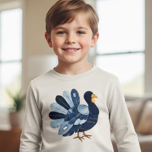Könnte beinhalten: Ein cremefarbenes Langarmshirt mit einem Truthahnmotiv aus Jeansstoff-Flicken. Der Truthahn hat einen dunkelblauen Körper, einen gelben Schnabel und Jeansfedern. Das Shirt wird von einem jungen Kind getragen.