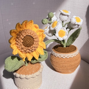 Puede incluir: Dos plantas en maceta de ganchillo. Un girasol amarillo con hojas verdes en una maceta de color crema con base marrón. Un ramo de flores de ganchillo blancas con centros amarillos en una maceta marrón con ribete de encaje blanco.
