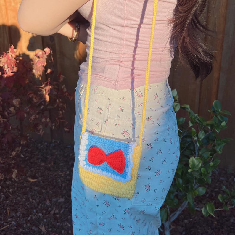 May include: A yellow, blue, and white crocheted bag with a red bow. The bag is worn as a crossbody purse.