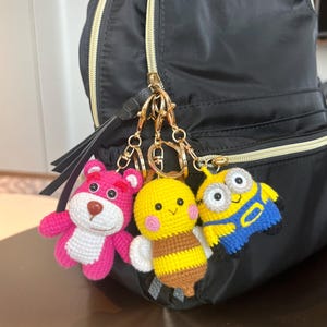 May include: A black backpack with three crocheted keychains attached to the zipper. The keychains are a pink bear, a yellow bee, and a blue minion.