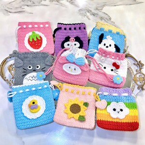 May include: A collection of handmade crochet drawstring pouches in various colors and designs. Each pouch features a different character or motif, including a strawberry, a cat, a duck, a sunflower, and a cloud. The pouches are displayed on a decorative tray.
