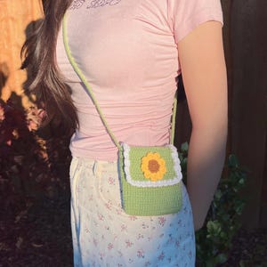 May include: A green crocheted crossbody bag with a white trim and a yellow sunflower design. The bag is worn over a pink shirt and a white floral skirt.