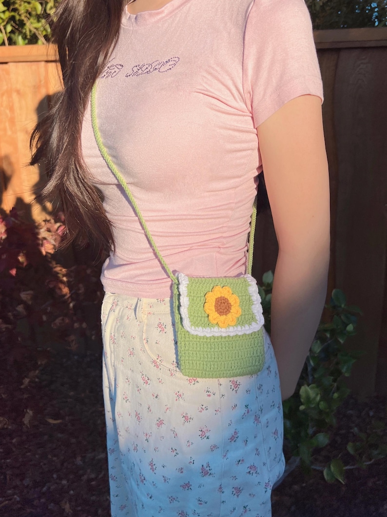 May include: A green crocheted crossbody bag with a white trim and a yellow sunflower design. The bag is worn over a pink shirt and a white floral skirt.