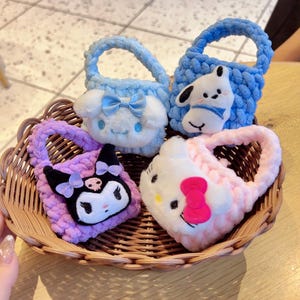 May include: Four plush, small tote bags in a woven basket. The bags are light blue, purple, and pink, each with a character face. One bag has a white dog, another has a white cat with a pink bow, a third has a purple cat, and the last has a white cat with a blue bow.