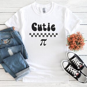 May include: White t-shirt with a black graphic that says "Cutie Pi" with a checkered pattern underneath the word "Cutie".