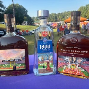 May include: Three bottles of liquor on a purple surface. The bottles are labeled "1800 Tequila" and "Woodford Reserve Kentucky Straight Bourbon Whiskey". Each bottle has a label depicting a football stadium. The background shows people and cars.