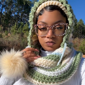 May include: A handmade crochet hood and cowl set in shades of green, cream, and white, with a fluffy pom-pom accent. The hood is designed with a matching cowl, creating a cozy and stylish accessory. The set is perfect for cold weather.