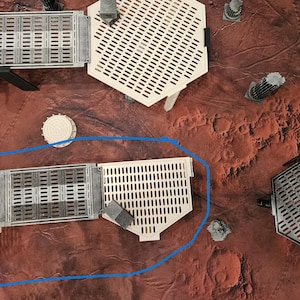 May include: An aerial view of a tabletop game setup featuring modular plastic terrain pieces in grey and white. The pieces include platforms, walkways, and structures, arranged on a reddish-brown surface. Small, grey square blocks are scattered around.