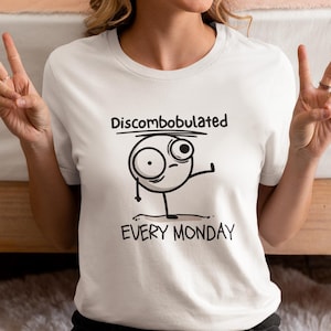 May include: White t-shirt with a black and white illustration of a stick figure with a confused expression. The text "Discombobulated Every Monday" is printed on the shirt.