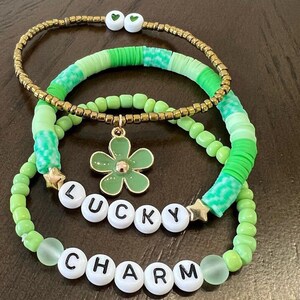 May include: A stack of four bracelets in shades of green and gold. One bracelet features the words "LUCKY CHARM" spelled out in white letter beads. Other bracelets have green beads, gold beads, and a green flower charm.