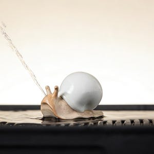 May include: A ceramic snail ornament with a white shell and a beige body, ejecting a stream of water. The snail sits on a dark, textured surface, with water around it. The background is a soft, gradient beige.