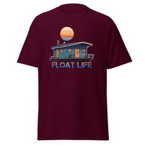 Puede incluir: Una camiseta de color granate con un gráfico de una casa flotante en el agua con una puesta de sol de fondo. El texto "FLOAT LIFE" está impreso debajo de la casa flotante.