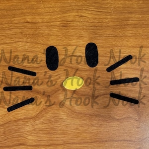 May include: A wooden surface with felt cutouts arranged to resemble a cat's face. The design includes two black ovals for eyes, a yellow oval for the nose, and black lines for whiskers. The text "Nana's Hook Nook" is visible.