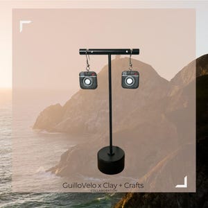 Puede incluir: Un par de pendientes negros con forma de cámara, colgados de un expositor negro. Los pendientes tienen un diseño detallado con una lente y un flash. El expositor tiene una base redonda y una barra horizontal. El texto "GuilloVelo x Clay + Crafts" es visible.