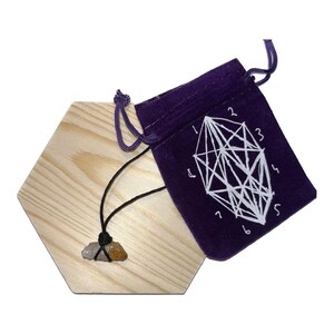 Puede incluir: Un collar con un colgante de piedra multicolor sobre un cordón negro, exhibido sobre una base hexagonal de madera clara. Una bolsa de terciopelo morado con un diseño geométrico blanco y números está al lado de la base.