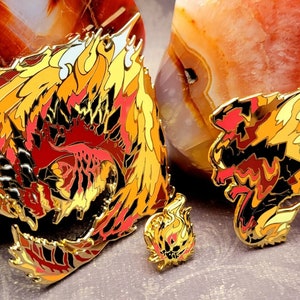 May include: Three gold-colored enamel pins featuring a fiery dragon design. The largest pin is a detailed depiction of a dragon with red, orange, and yellow flames. The medium-sized pin is a simplified version of the dragon with a similar color scheme. The smallest pin is a simple flame design.