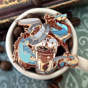 May include: A coffee-themed enamel pin featuring a dragon design. The pin depicts a coffee cup with a dragon, coffee beans, and a coffee maker. The dragon is in shades of blue, brown, and tan. The pin is set against a white coffee cup.