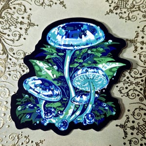 Pode incluir: Um autocolante com um grupo de cogumelos estilizados em tons de azul e verde. As tampas dos cogumelos têm um aspeto facetado, semelhante a uma gema, com gotas de água. O fundo é preto e o autocolante está numa superfície estampada.