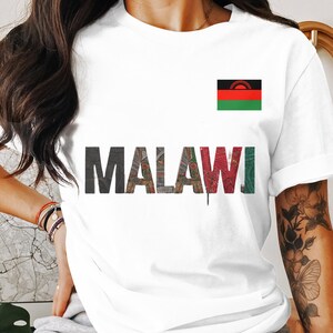 Puede incluir: Camiseta blanca con la palabra "MALAWI" en una fuente multitéxtil y la bandera de Malaui en el pecho. La bandera es un tricolor horizontal de negro, rojo y verde.