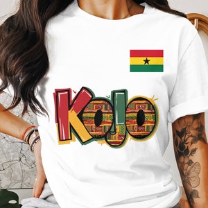 Pode incluir: Camiseta branca com a palavra "Kojo" em letras coloridas e estampadas. O design incorpora vermelho, verde, amarelo e preto. Uma pequena bandeira de Gana está no canto superior direito. A camiseta é provavelmente de algodão.
