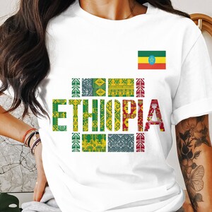 May include: White t-shirt featuring the word "ETHIOPIA" in colorful, patterned letters. The Ethiopian flag is displayed on the upper right. The shirt is a casual wear item.