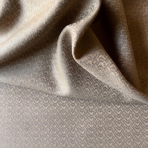 May include: A piece of taupe-colored fabric featuring a repeating, small-scale chevron or arrow-like pattern. The material is draped to show soft folds and highlights, revealing a consistent, textured weave throughout the entire surface area.