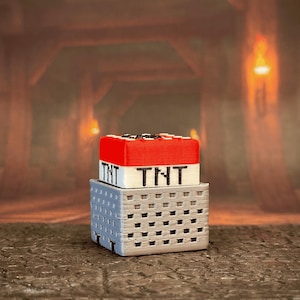 May include: A 3D-printed TNT block, featuring a red top with black detailing, sits atop a gray, perforated base. The word "TNT" is printed in black on a white background. The item is designed to resemble a block from a popular video game.