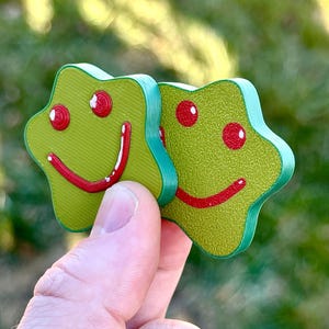 Puede incluir: Dos adornos de madera verdes en forma de estrella con ojos rojos y sonrisas rojas. Los adornos tienen una superficie texturizada y un borde turquesa. Los adornos están sostenidos en la mano de una persona.