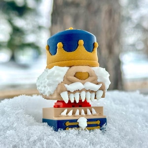 Puede incluir: Una figura de cascanueces impresa en 3D con una corona azul y dorada, barba blanca y dientes grandes. La figura tiene una base roja y se asienta sobre una plataforma azul y dorada. El fondo está borroso con nieve.