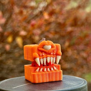 May include: A 3D-printed orange monster figure with sharp white teeth and a single eye. The figure has a boxy shape and is set against a blurred background of fall foliage. The figure is a unique decorative item.