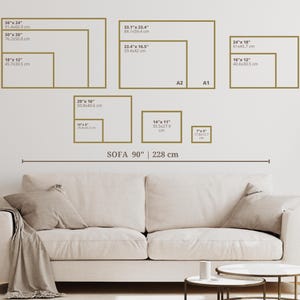 Op de afbeelding: Een witte bank met een bruin wollen deken erop. De bank is 90 inch (228 cm) lang. Aan de muur achter de bank hangen verschillende ingelijste foto's. De foto's zijn van verschillende grootte en zijn in een rasterpatroon gerangschikt. De foto's zijn allemaal in gouden lijsten. De tekst op de foto's geeft de grootte van de foto's in inches en centimeters aan.