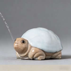 May include: A ceramic turtle figurine with a light blue shell and a tan body. A thin stream of water is coming from the turtle's mouth. The figurine is on a gray background.