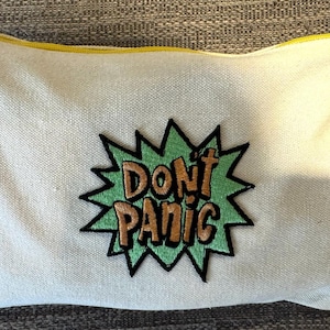 Op de afbeelding: Witte canvas toilettas met een groene en oranje geborduurde patch met de tekst "Don't Panic".