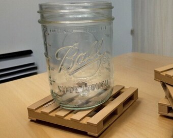 May include: A clear glass jar with the text "Ball Wide Mouth" on the side, sitting on a small wooden pallet coaster. There are three additional wooden pallet coasters stacked on top of each other in the background.