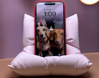 May include: A red iPhone with a white screen displaying the time 1:09 and the date Wednesday, June 5. The phone is propped up in a white pillow with two brown and black dachshund dogs on the screen.
