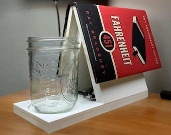May include: White book stand with a red and black book titled "Fahrenheit 451" by Ray Bradbury. A clear glass jar is sitting on the stand.