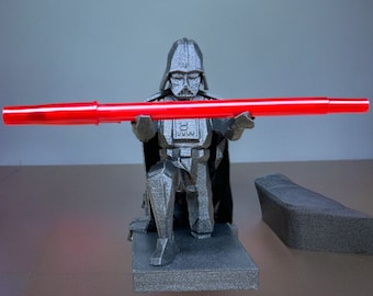 Puede incluir: Una figura de Darth Vader impresa en 3D de color negro sosteniendo un sable de luz rojo. La figura está arrodillada sobre una base negra.