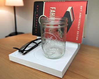 May include: White organizer with a book titled "Fahrenheit 451" by Ray Bradbury, a glass jar, and a pair of black glasses.