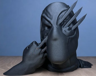 Puede incluir: Una figura impresa en 3D de color negro de una figura enmascarada con cuchillas afiladas que sobresalen de su ojo. La figura tiene la mano levantada hacia su boca, como si estuviera haciendo callar a alguien.