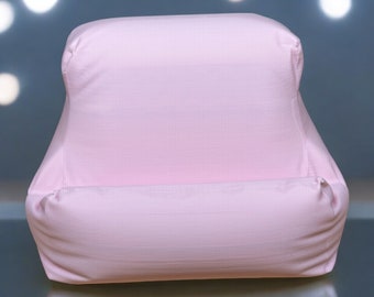 Puede incluir: Una almohada rectangular de color rosa claro con una parte superior curvada y un pequeño labio en la parte inferior. La almohada está hecha de una tela suave y texturizada. El fondo es un degradado de azul y gris con luces blancas borrosas.