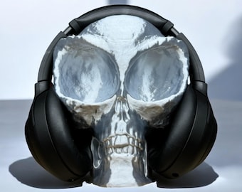 Puede incluir: Auriculares negros de diadema con un soporte en forma de cráneo. El cráneo es blanco con detalles grises.