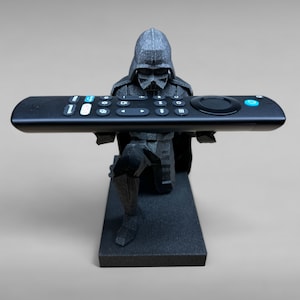 May include: A black Darth Vader figurine holding a black remote control with a blue button. The figurine is standing on a black base.