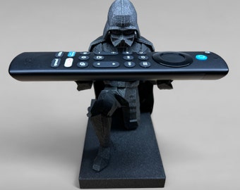 Puede incluir: Una figura negra de Darth Vader sosteniendo un mando a distancia negro con un botón azul. La figura está de pie sobre una base negra.