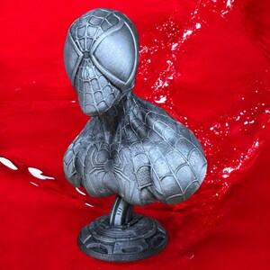 May include: A silver-colored bust of Spider-Man, featuring intricate web detailing across the mask and suit. The bust is mounted on a circular base and set against a red background.