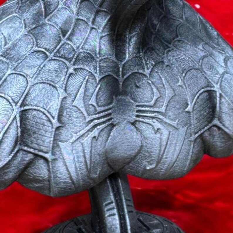 May include: A gray sculpture featuring a spiderweb design and a spider emblem. The sculpture has a textured surface and is mounted on a black base. The overall design suggests a superhero theme.