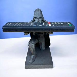 May include: A black 3D printed Darth Vader figurine holding a black remote control in its hands. The figurine is kneeling on a black base.